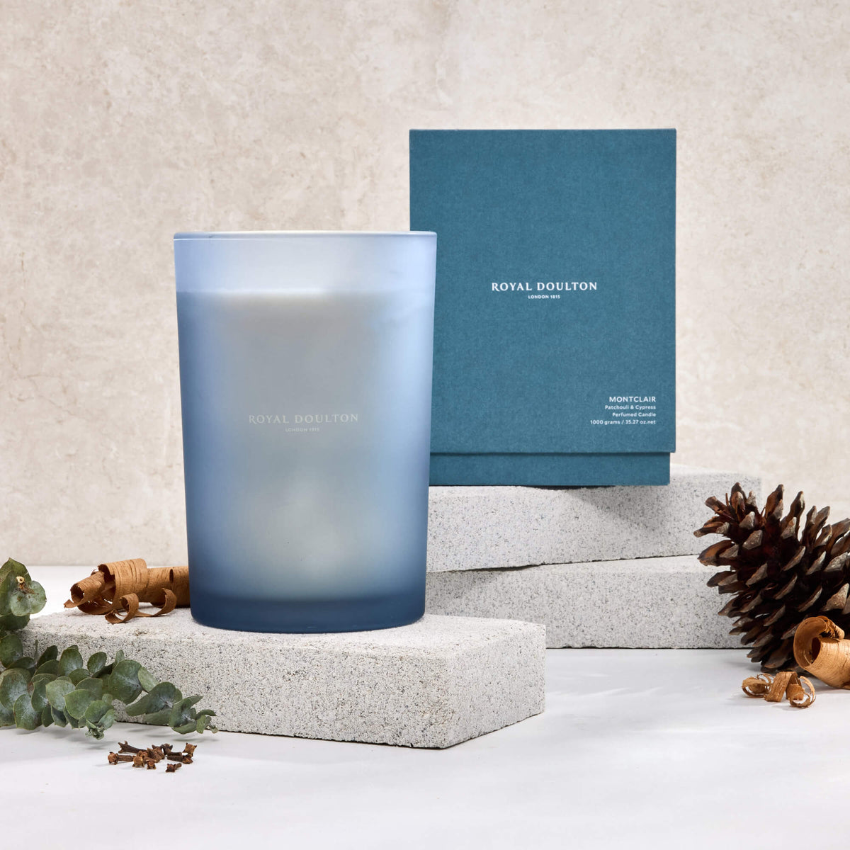 Royal Doulton Montclair 1000g candle in a tall blue vessel with its packaging box, pine cone and foliage on pale blocks.