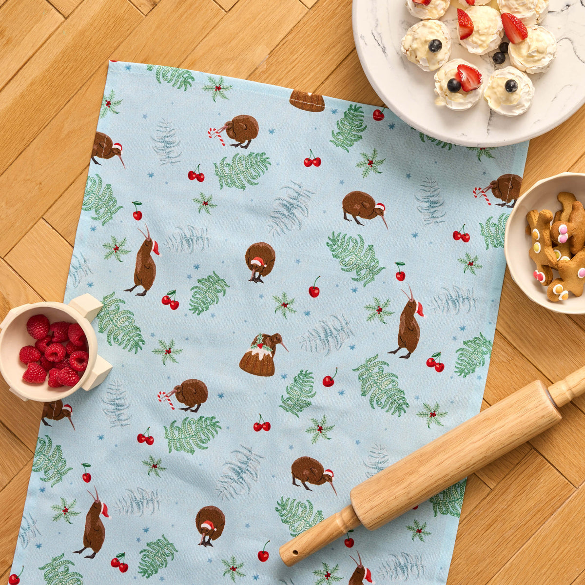 Pale blue Twill & Co Kiwiana Christmas tea towel with kiwi birds, foliage and cherries beside a rolling pin