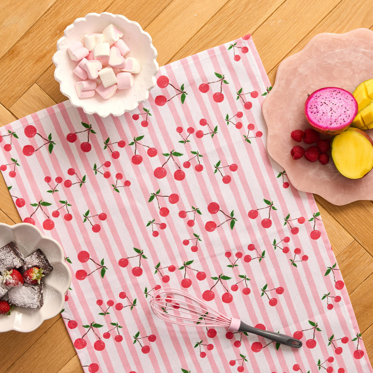 Pink striped Twill & Co cherry-print tea towel laid beneath a whisk and bowls of fruit and sweets
