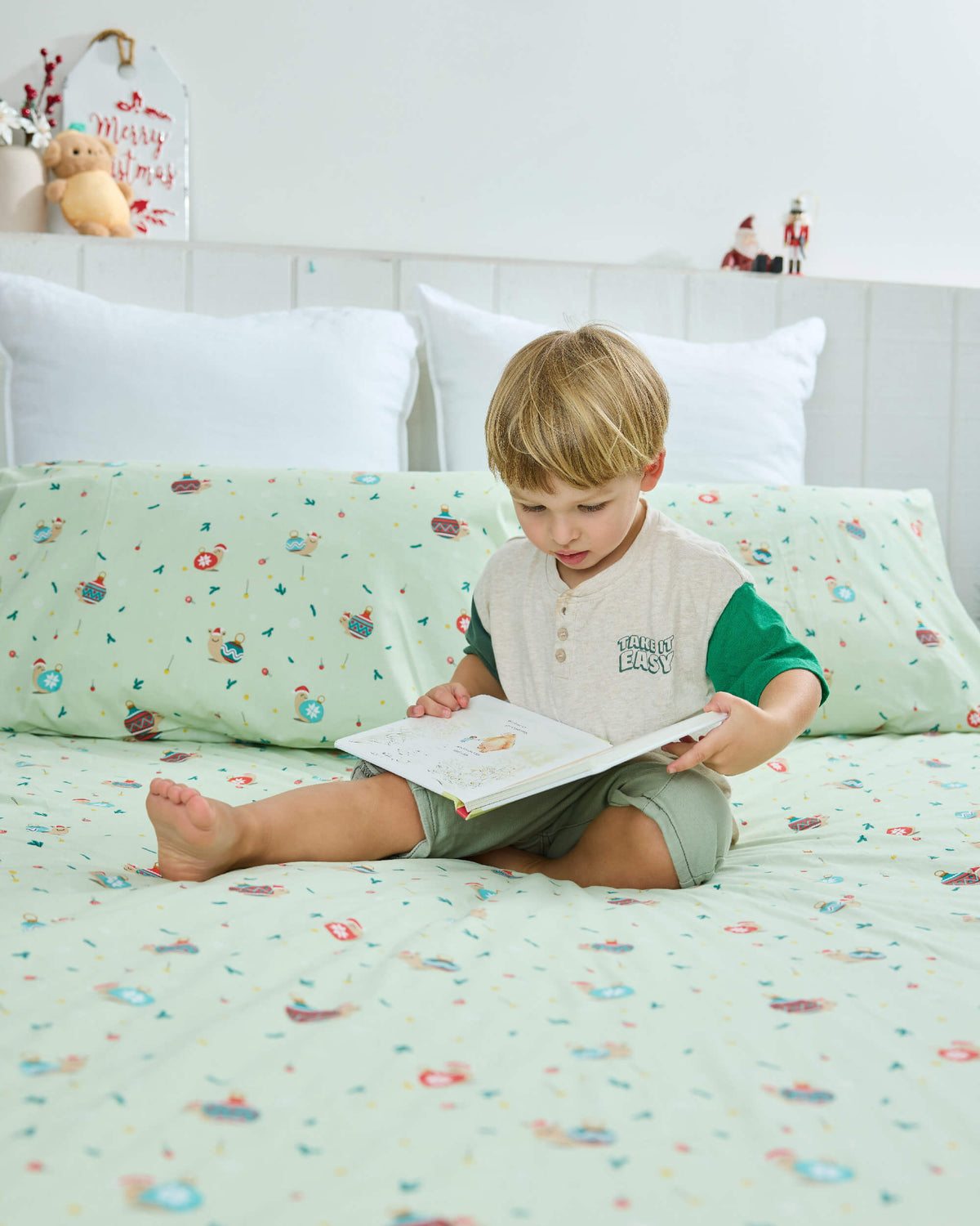 Child reading cross-legged on mint-green Twill & Co Snail Baubles bedding beside a small Christmas tree