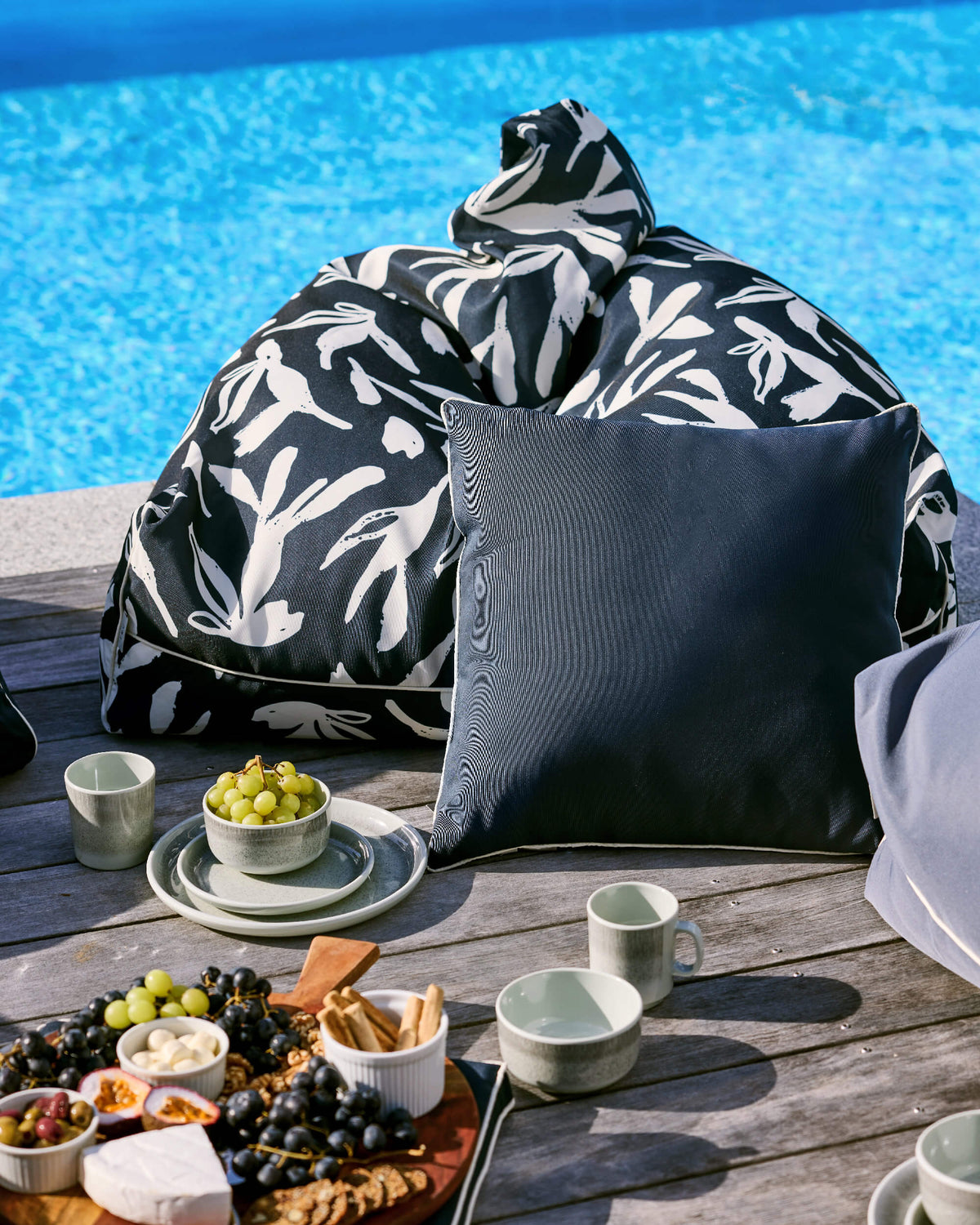 Black-and-white leaf-print Twill & Co bean bag behind a plain charcoal cushion at a poolside picnic