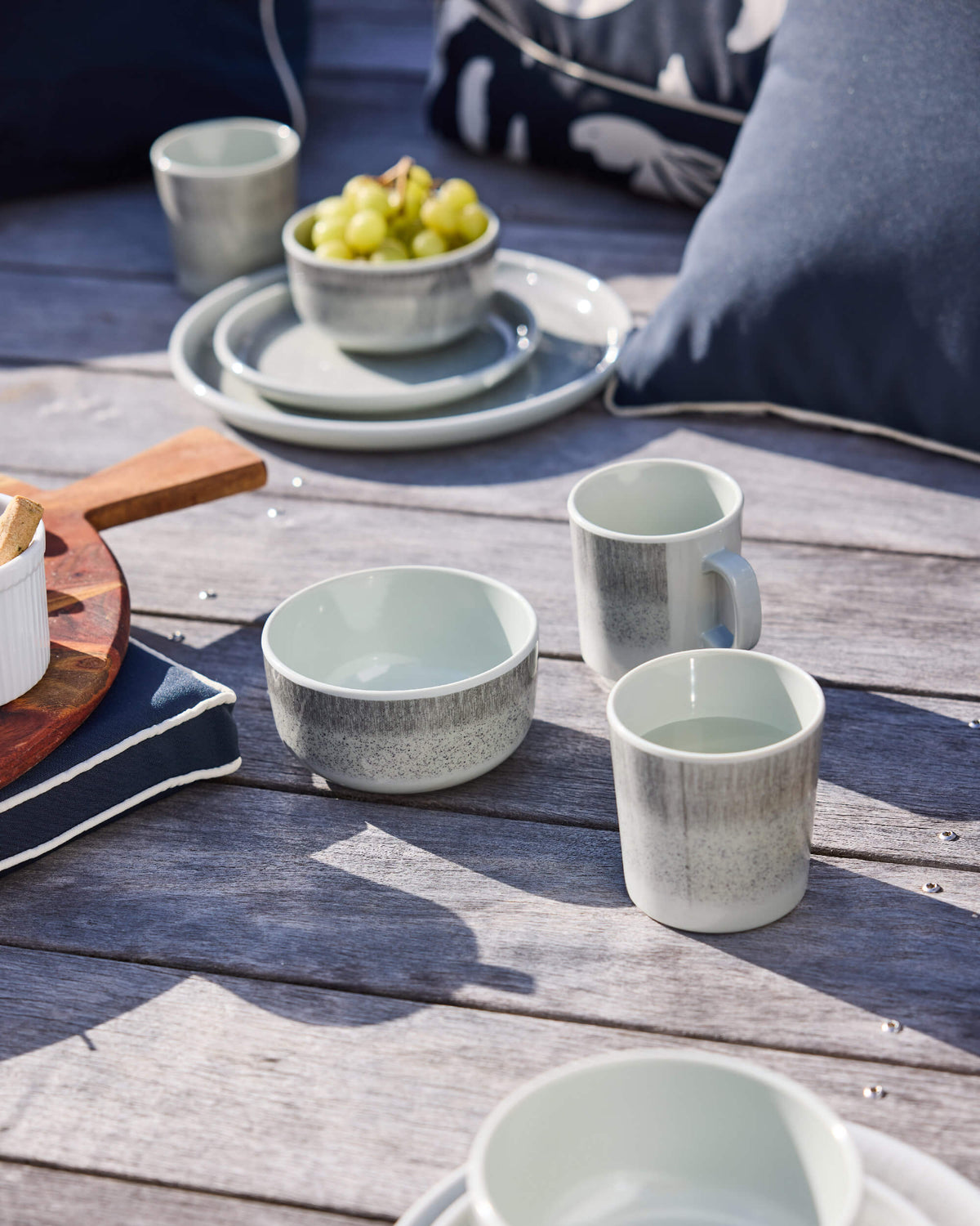 Pale sage Twill & Co mugs, tumblers and bowls on a timber deck beside charcoal cushions