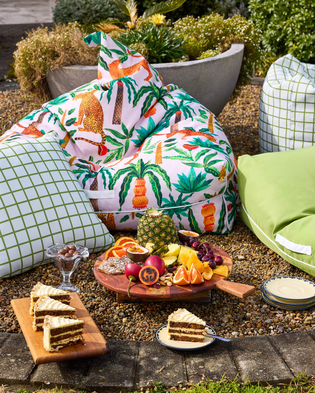 Leopard-print Twill & Co seating with green checked cushions beside fruit and cake on a gravel patio