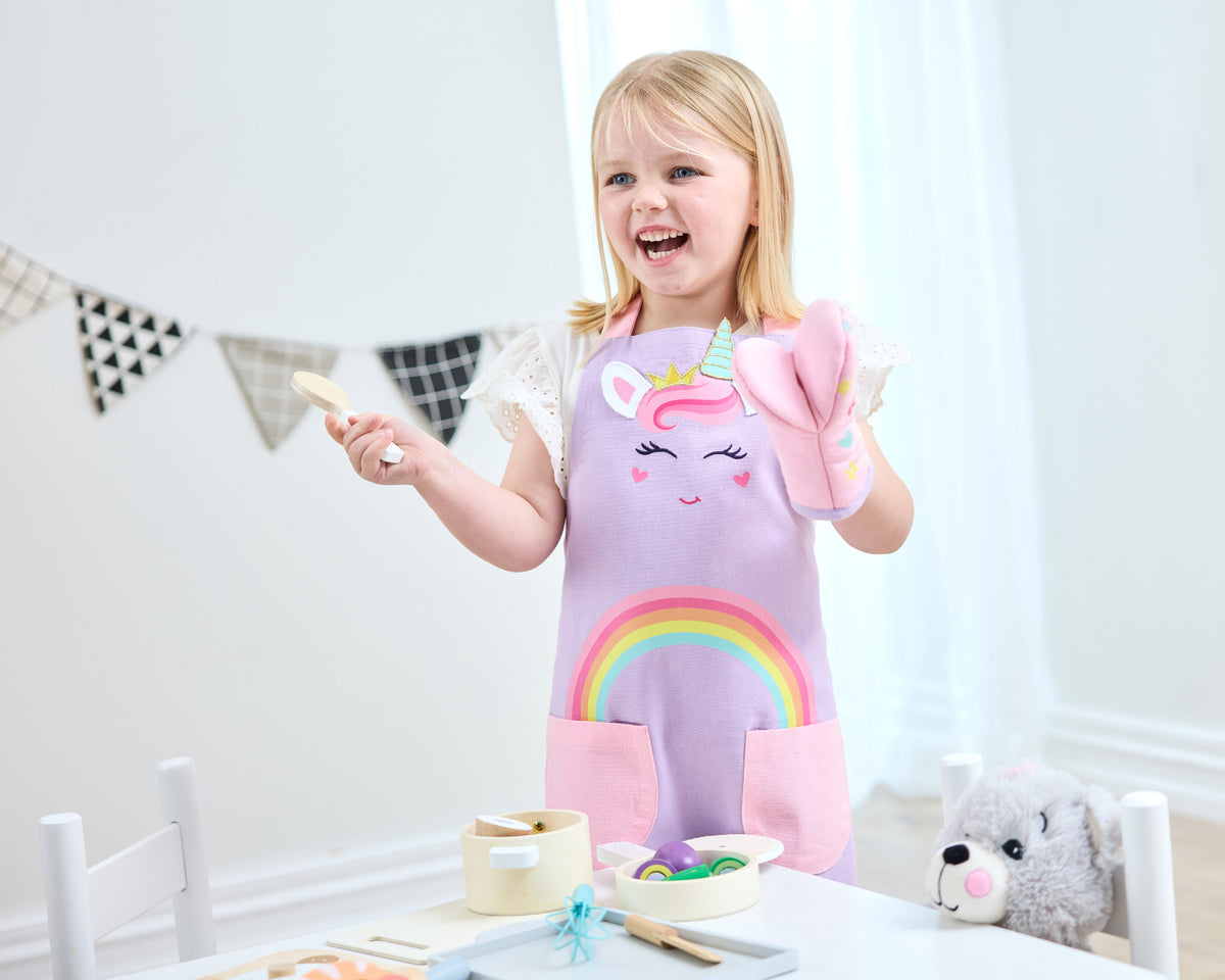 Child in a pink Twill & Co unicorn apron holding a utensil and matching oven mitt at a play table