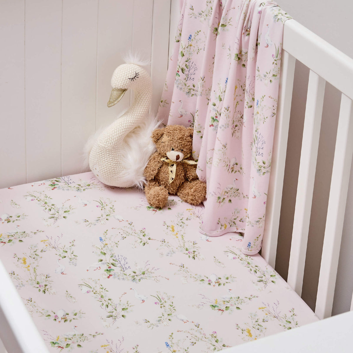 White cot with pink floral Twill & Co bedding, a draped wrap and swan and teddy bear toys