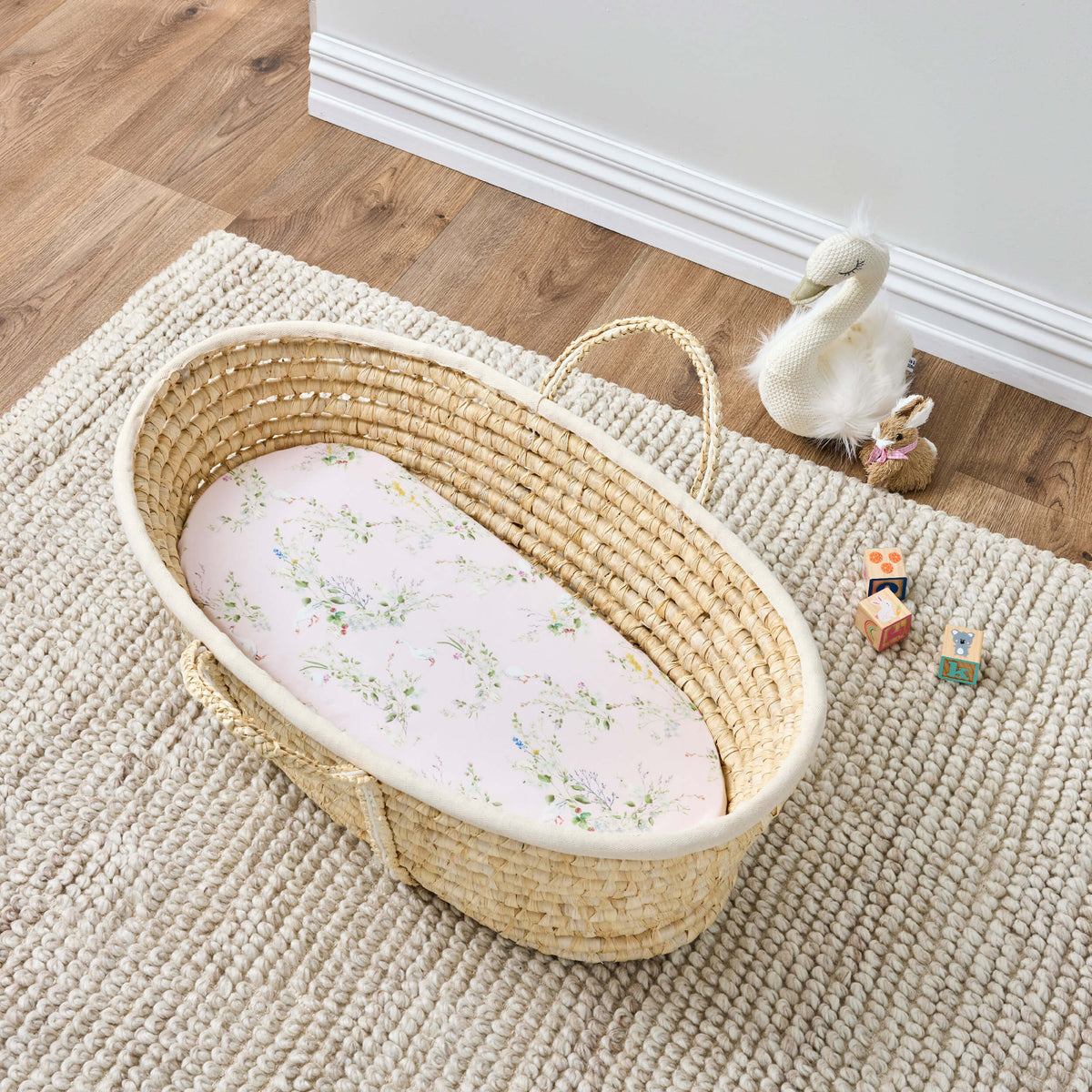 Overhead view of a woven bassinet with a pink floral Twill & Co sheet on a cream rug
