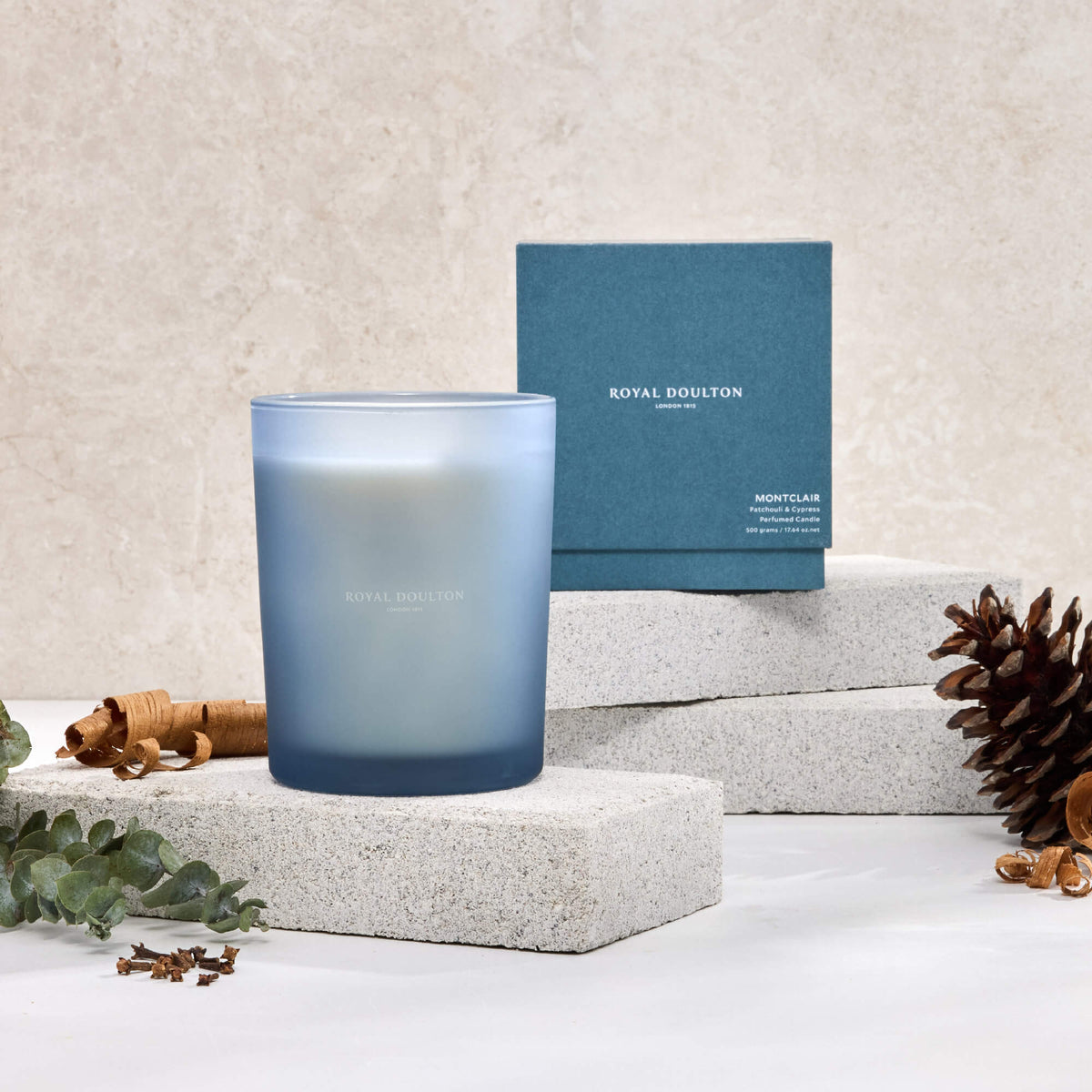 Royal Doulton Montclair 500g candle in a blue vessel beside its blue packaging box, with a pine cone and wood curls.