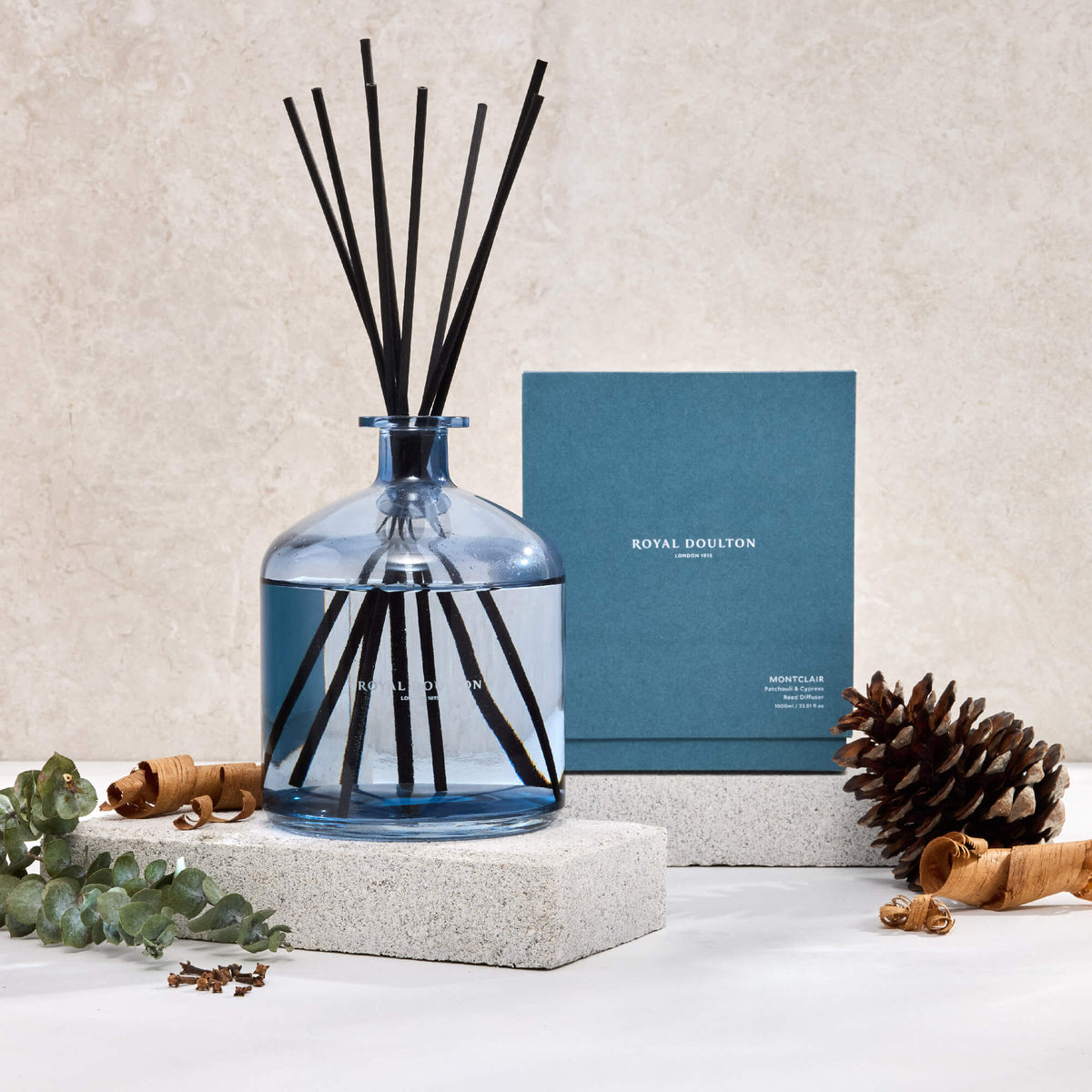 Royal Doulton Montclair 1000ml diffuser and blue packaging box on pale blocks, with black reeds, a pine cone and wood curls.