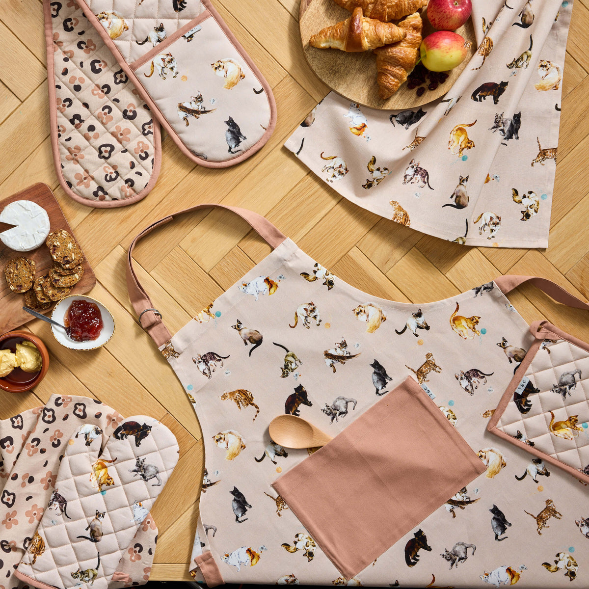 Pale pink Twill & Co cat-print apron and quilted oven gloves laid out with croissants and jam