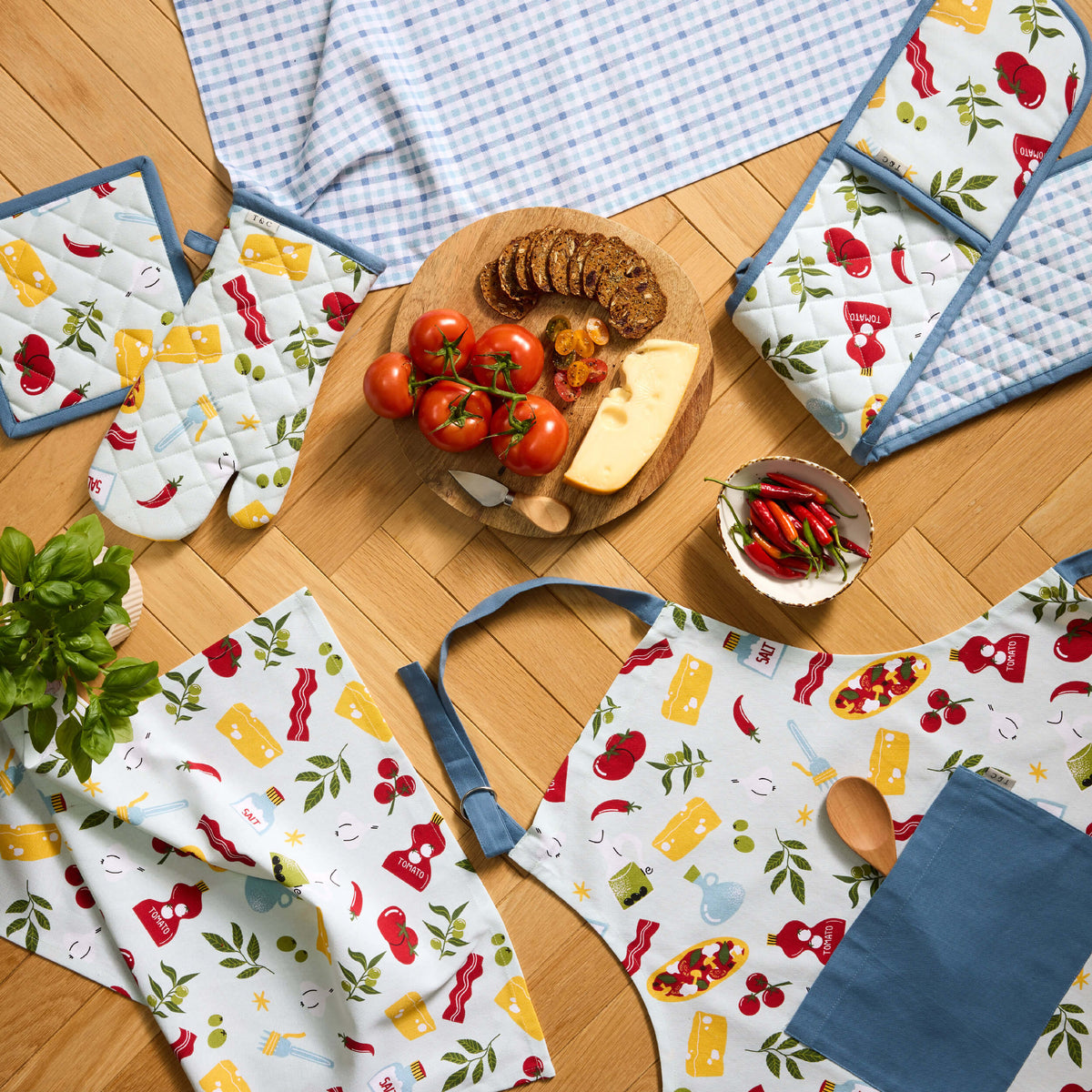 Twill & Co Picnic apron and oven gloves with colourful food motifs beside a blue checked tea towel