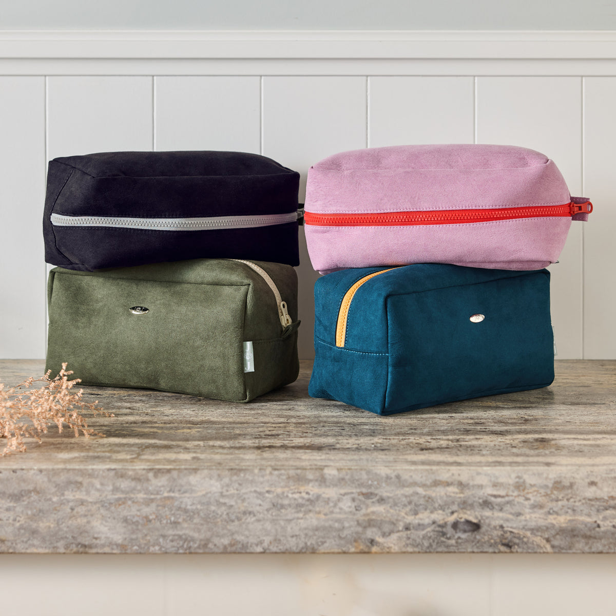 June & Jupiter washbags in black, pink, olive and teal stacked on a stone ledge