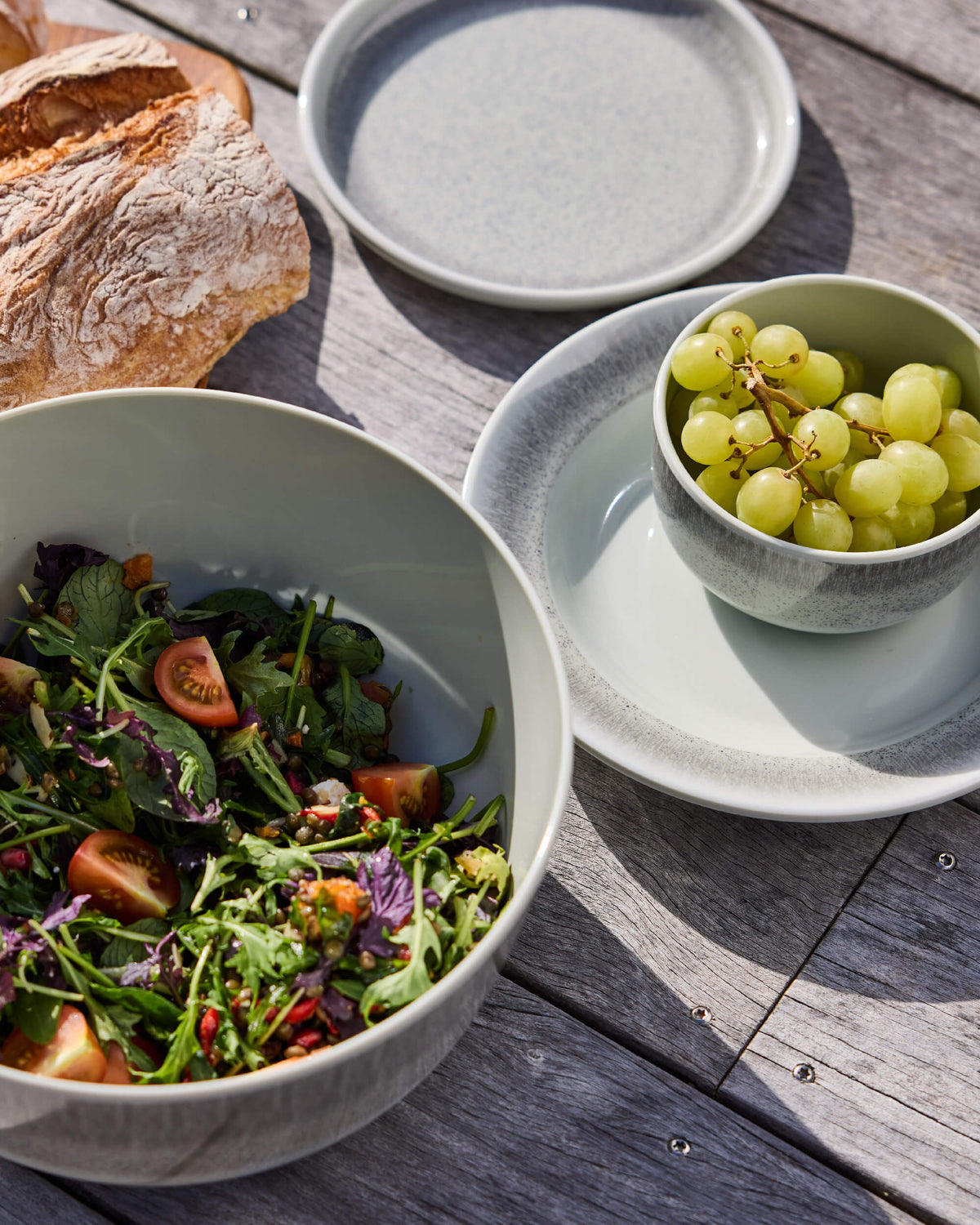 Overhead detail of pale sage Twill & Co tableware with a salad bowl, grapes and bread on weathered decking