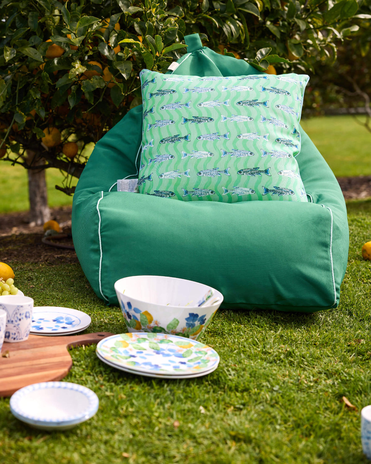 Plain green Twill & Co bean bag with a fish-print cushion beside patterned picnic plates on the lawn