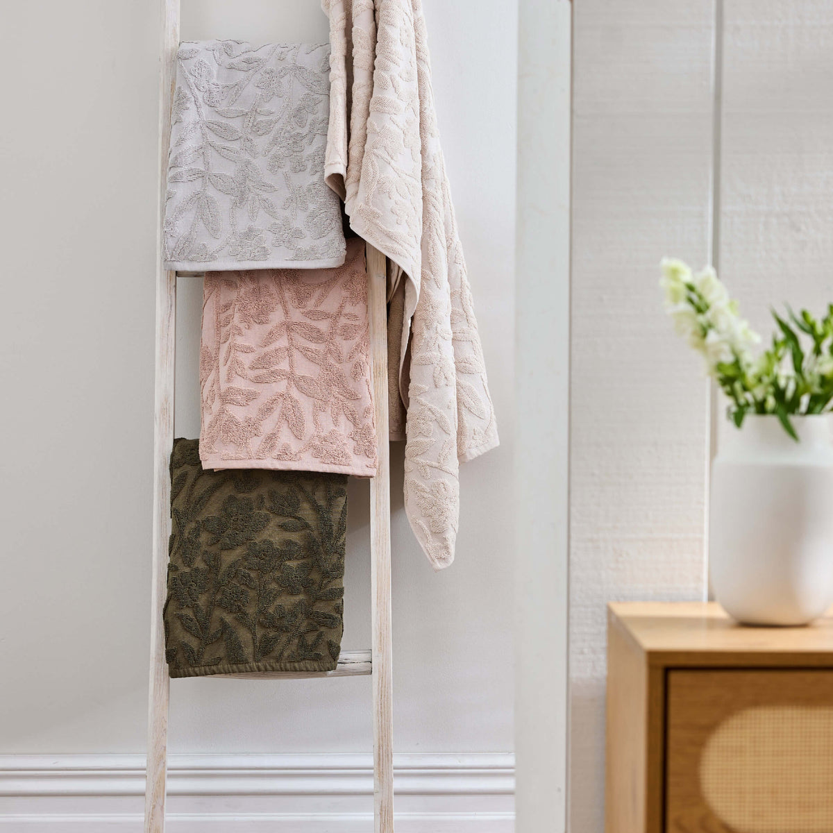 June & Jupiter Nova leaf-patterned towels in grey, pink, olive and pale neutral shades on a wooden ladder