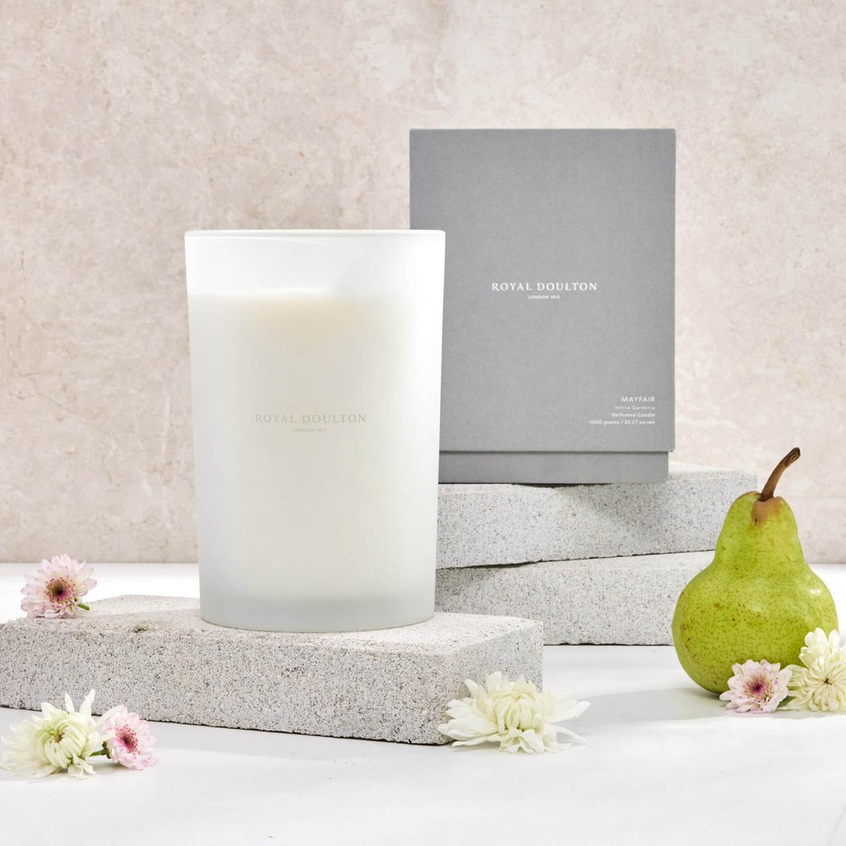 Royal Doulton Mayfair 1000g candle in a tall white vessel beside its grey packaging box, with a pear and flowers.